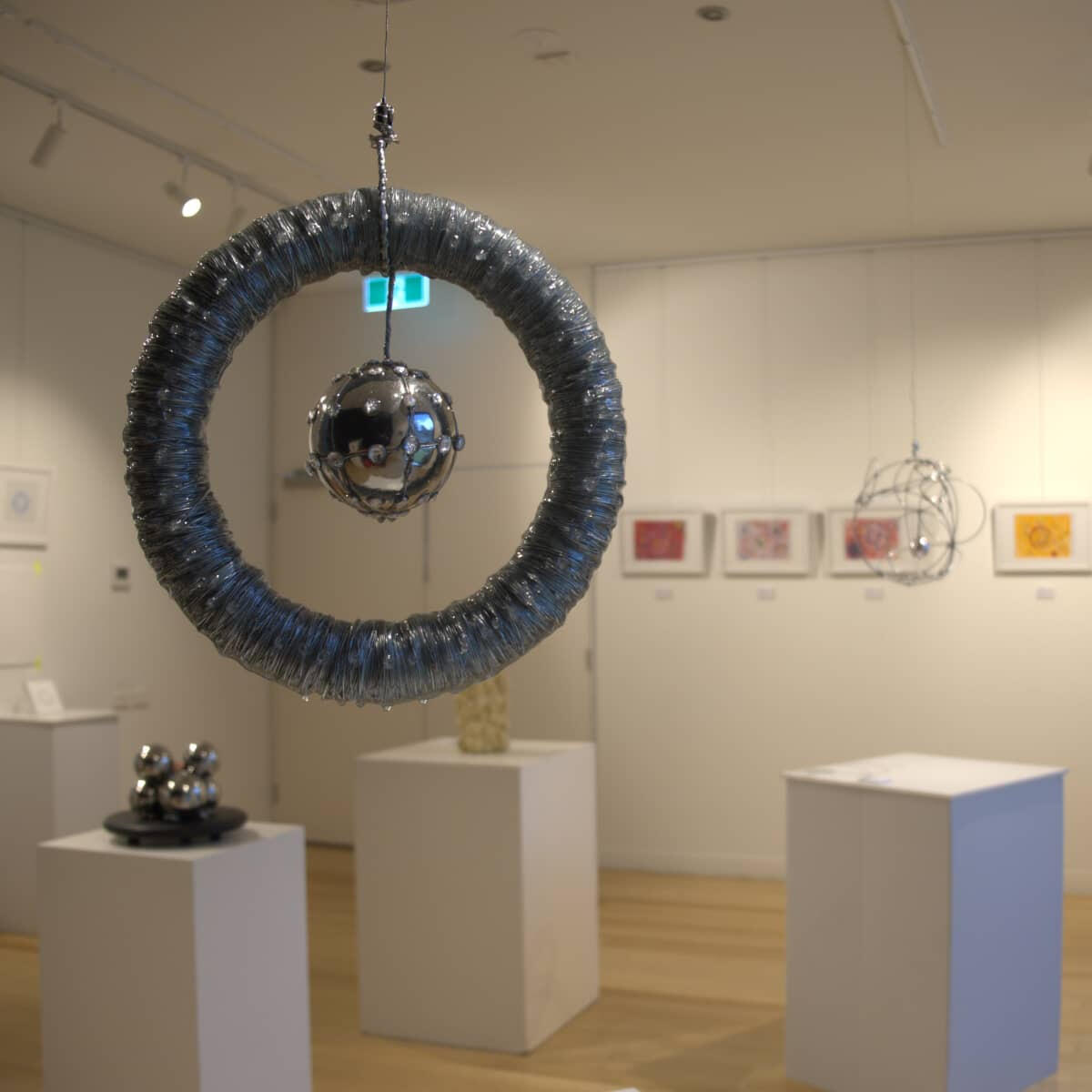 Michal Plis Contemporary Art Exhibition Photograph of a gallery exhibition featuring original contemporary artworks and sculptures by Melbourne abstract artist Michal Plis. A prominent, textured circular hanging sculpture is displayed in the foreground.